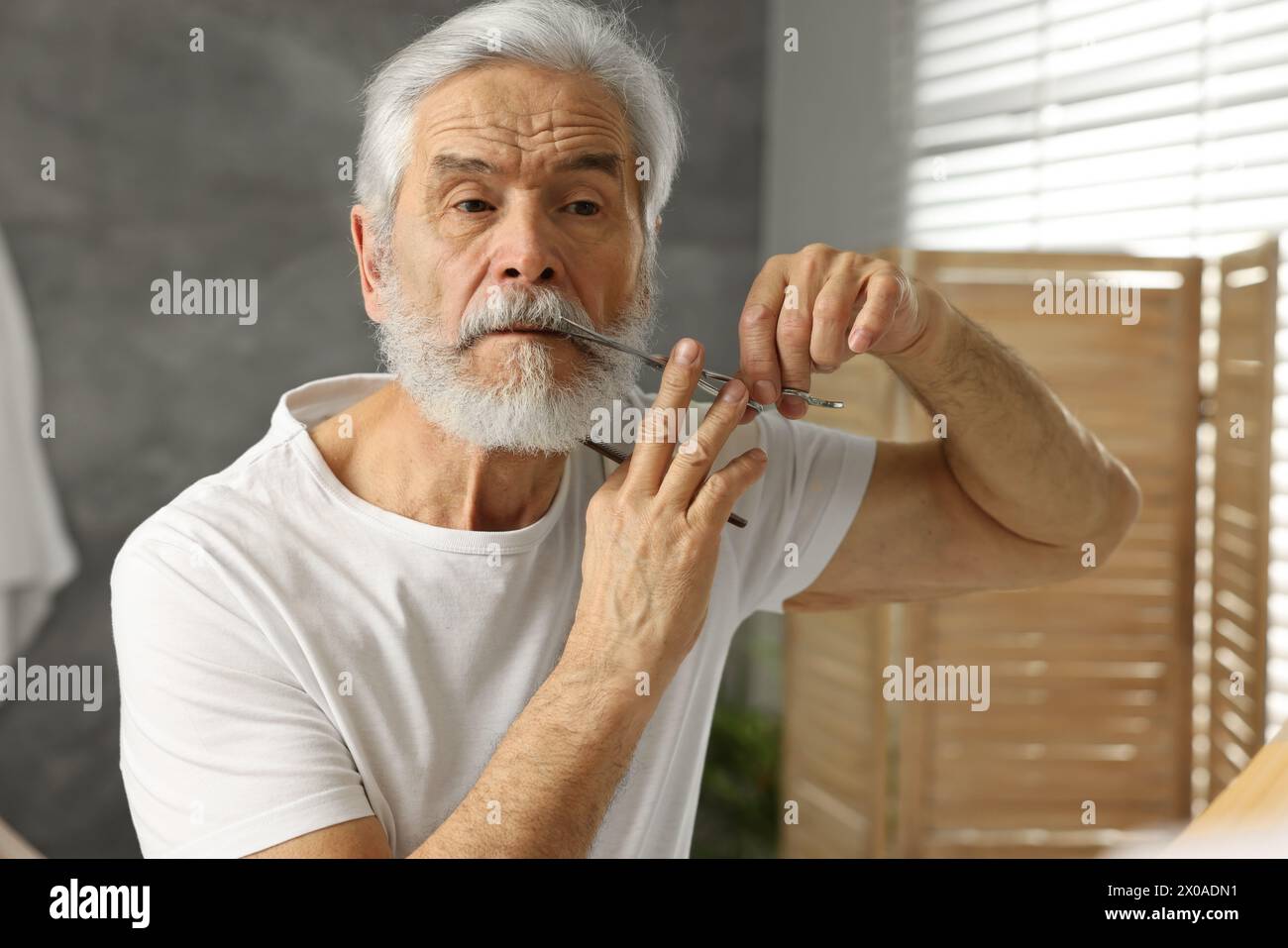 Senior man trimming mustache with scissors near mirror in bathroom