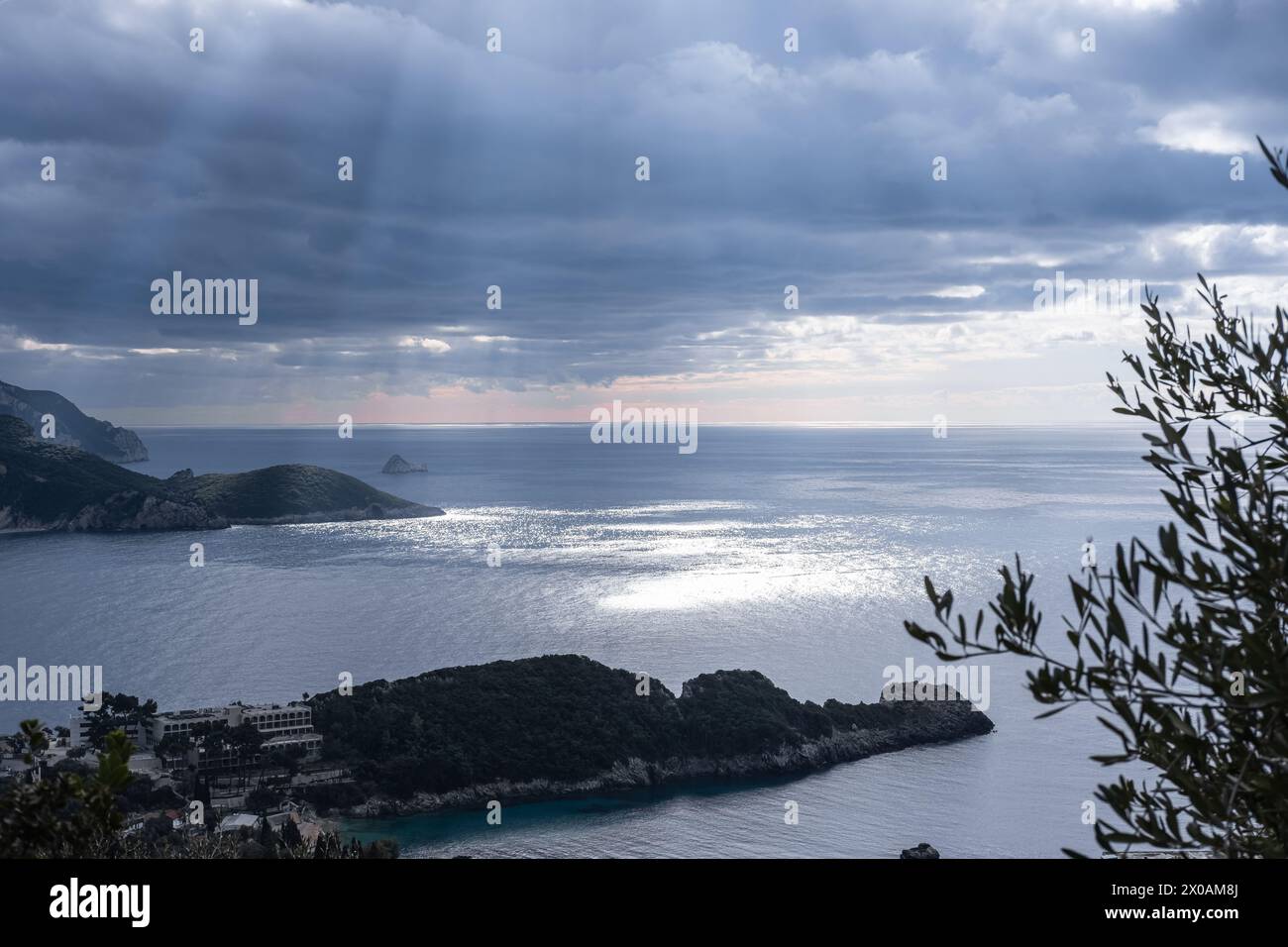 Natural light rays and blue cloudy sky over sea. Sunbeams reflections on calm sea water Corfu Greece. Seashore with a rays of sun against a moody dram Stock Photo