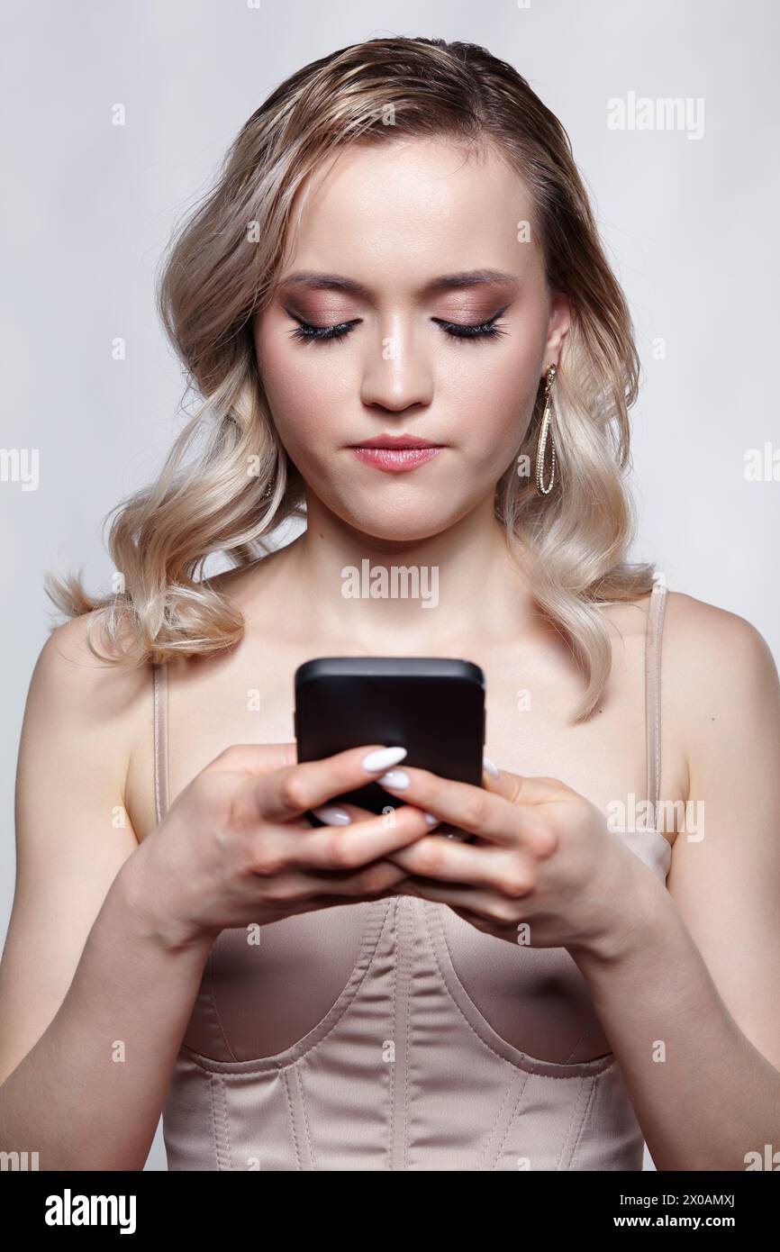 Portrait of young woman in beige dress on gray background. Female with a phone in hands typing SMS. Stock Photo