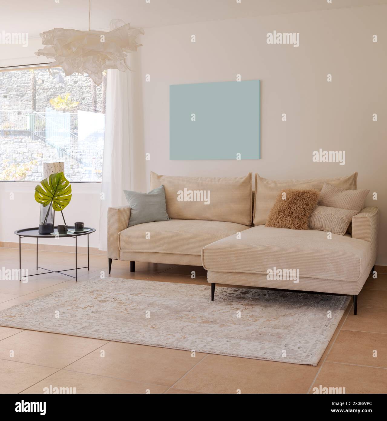 Detail of the living room with a light fabric sofa with cushions and a small table with plants next to it. Above the sofa there is space for a table. Stock Photo