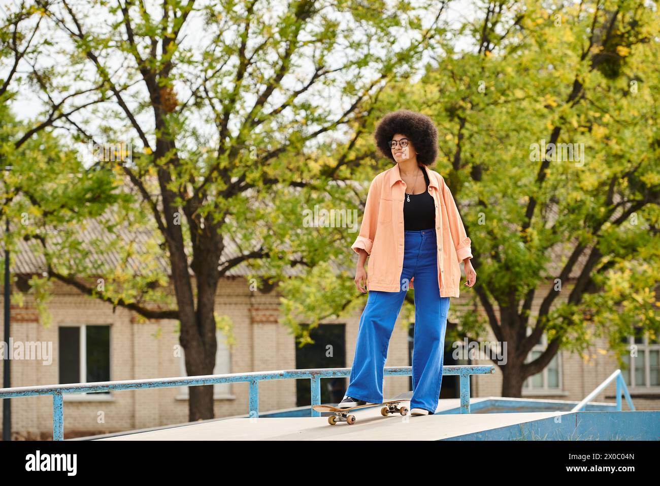 Young African American woman with curly hair skillfully riding a skateboard on top of a ramp at an outdoor skate park. Stock Photo