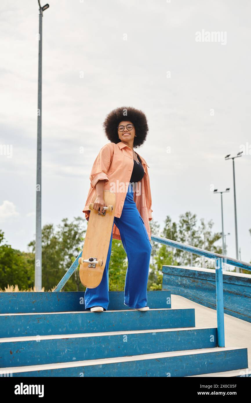 A young African American woman with curly hair confidently stands on steps, holding a skateboard in an urban skate park. Stock Photo