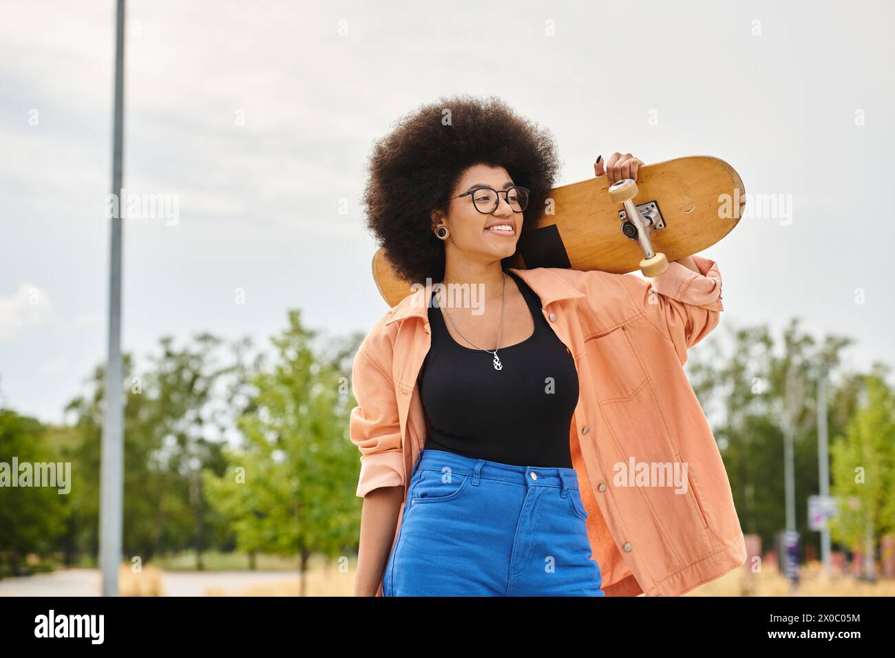 A young African American woman with curly hair holds a skateboard up to her face in a skate park. Stock Photo