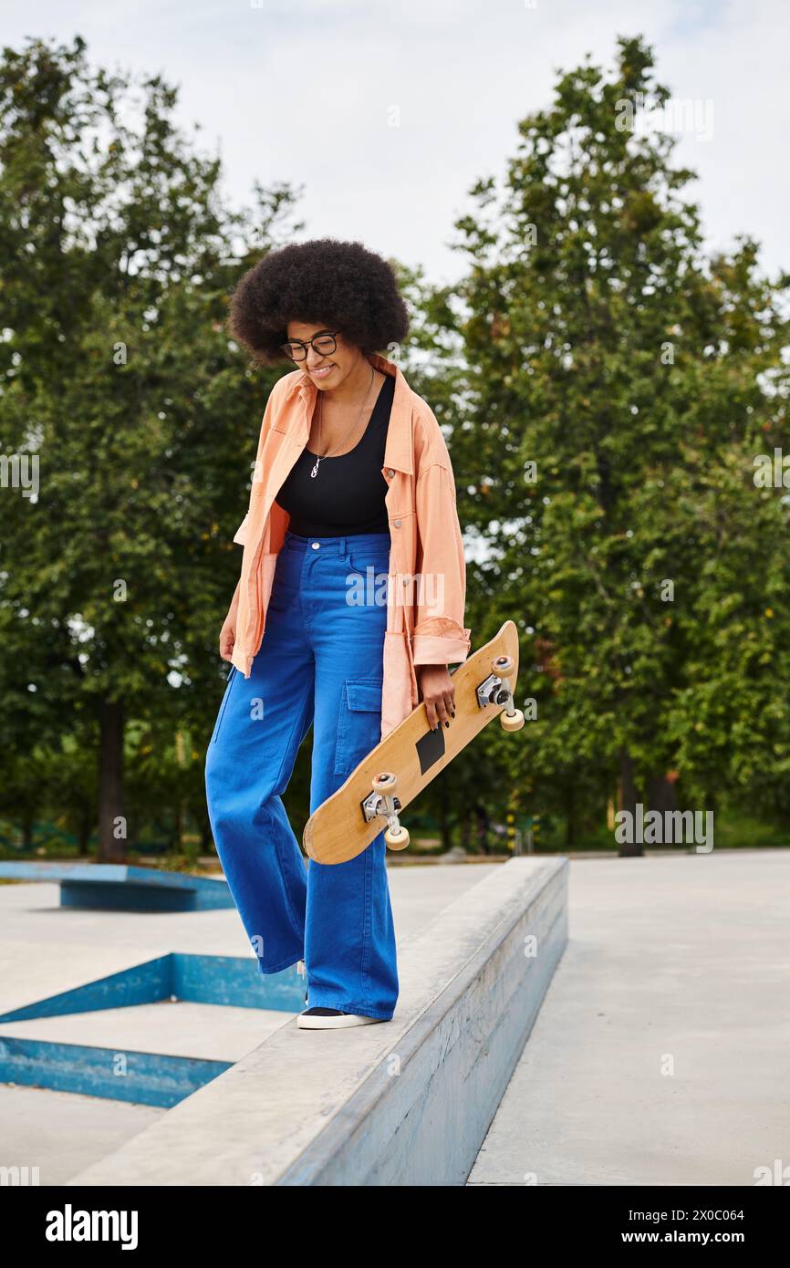 A young African American woman with curly hair stands confidently on a ledge with her skateboard in a skate park. Stock Photo