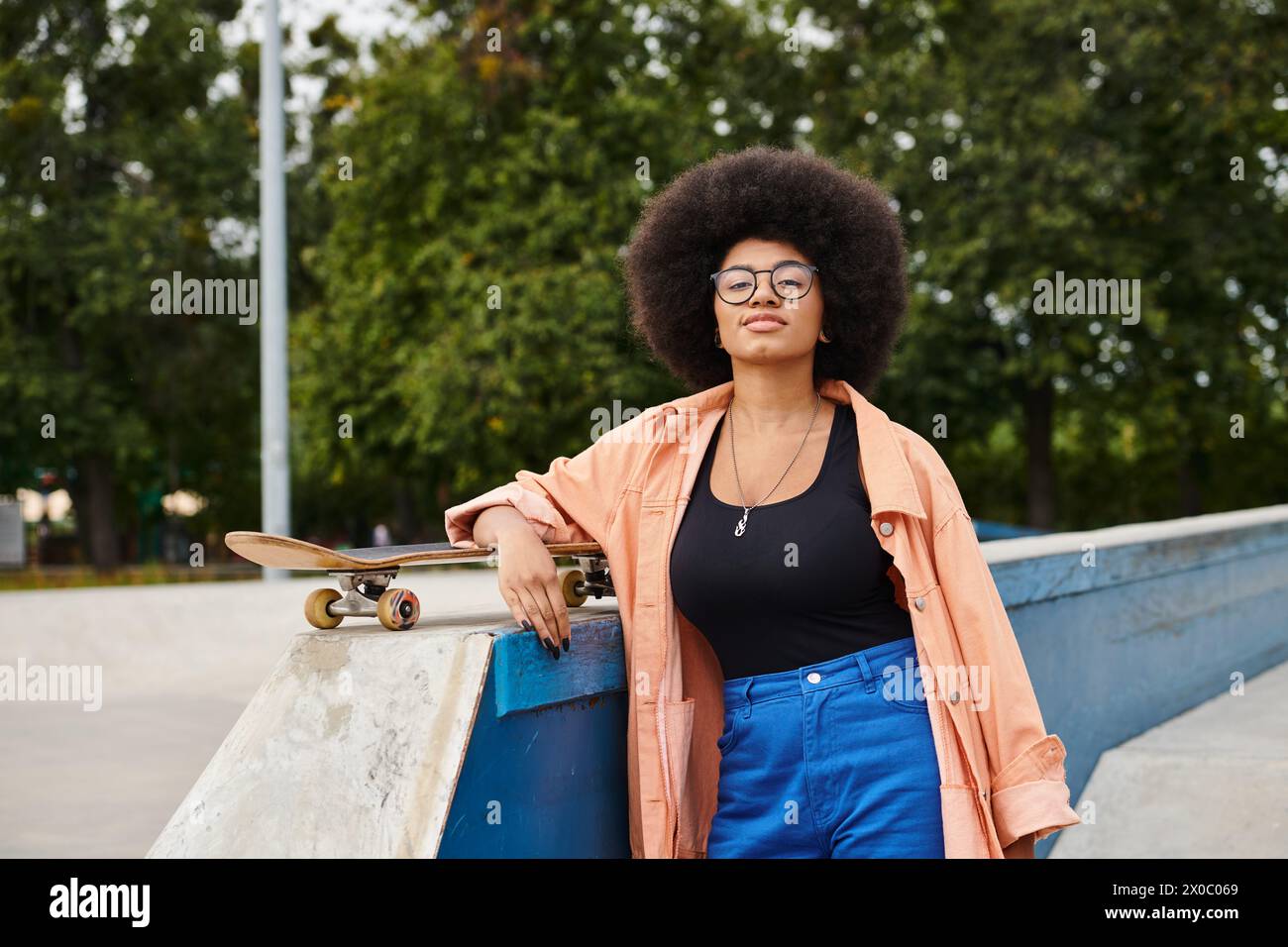 A young African American woman with curly hair stands confidently next to a skateboard on a skate park ramp. Stock Photo