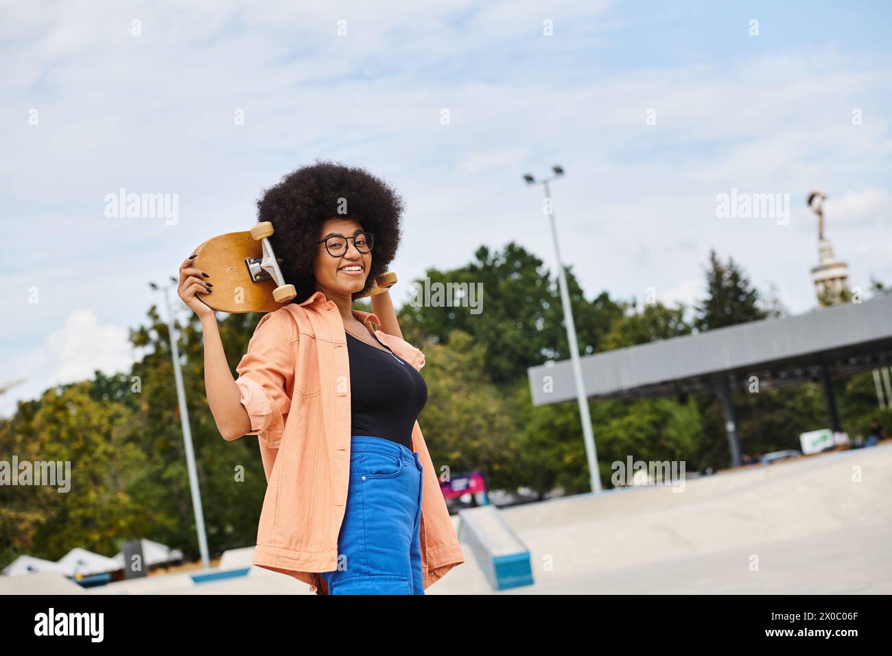 A stylish African American woman with curly hair holds a skateboard in her right hand at a skate park. Stock Photo