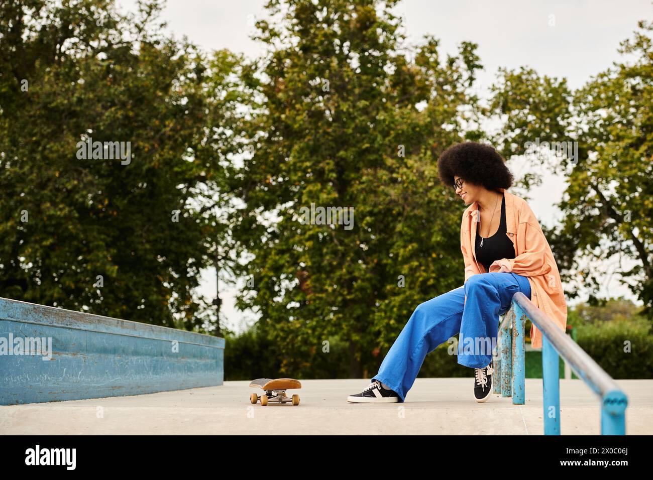 An African American woman with curly hair is sitting on a rail next to a skateboard in a skate park. Stock Photo