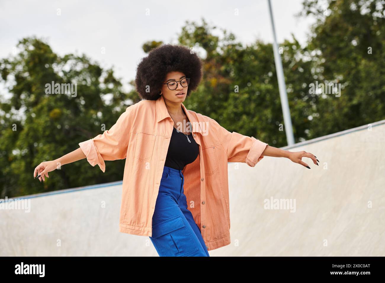 A young African American woman with curly hair confidently rides her skateboard up the side of a ramp at an outdoor skate park. Stock Photo