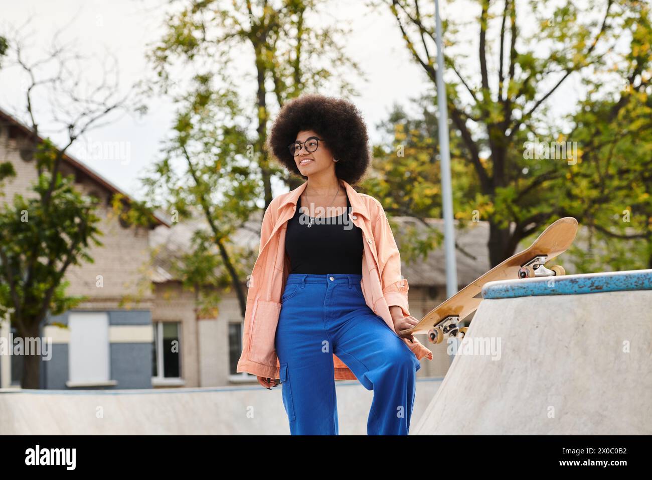 Young African American woman with curly hair stands next to skateboard on a skateboard ramp in a vibrant outdoor skate park. Stock Photo
