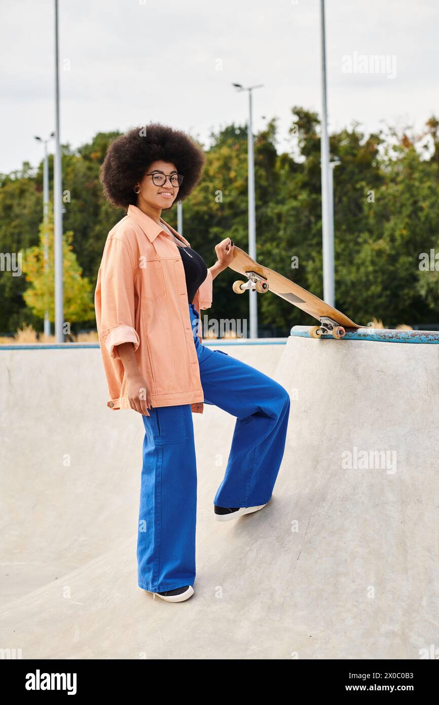 A young African American woman with curly hair skillfully standing on top of a skateboard ramp in an outdoor skate park. Stock Photo