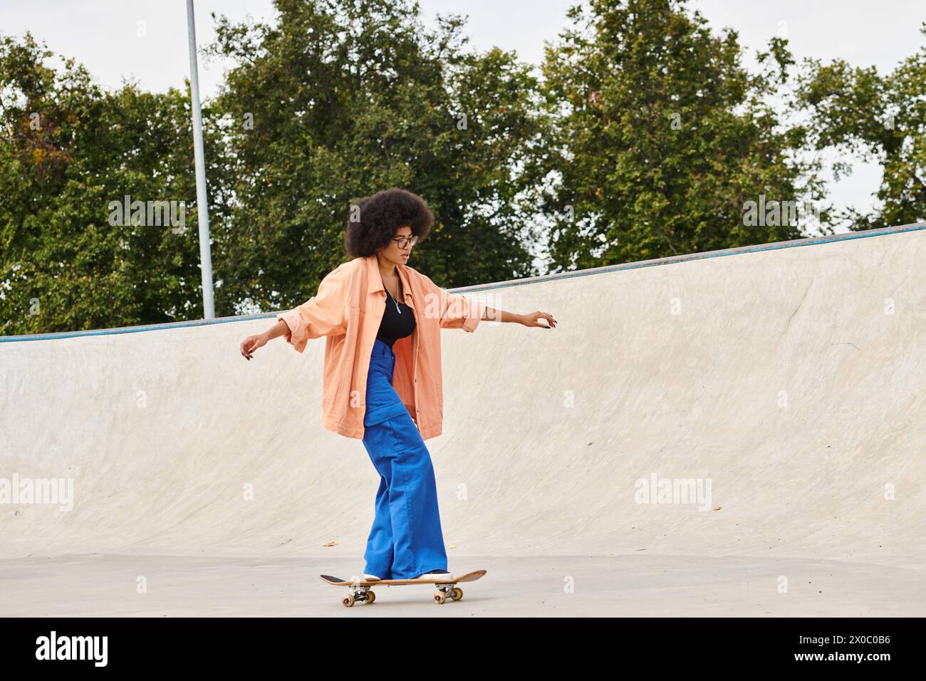 A young African American woman with curly hair skillfully rides a skateboard up the side of a ramp at an outdoor skate park. Stock Photo