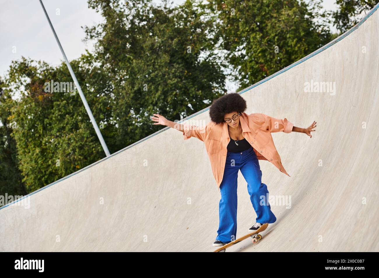 A young African American woman with curly hair riding a skateboard up the side of a ramp at an outdoor skate park. Stock Photo