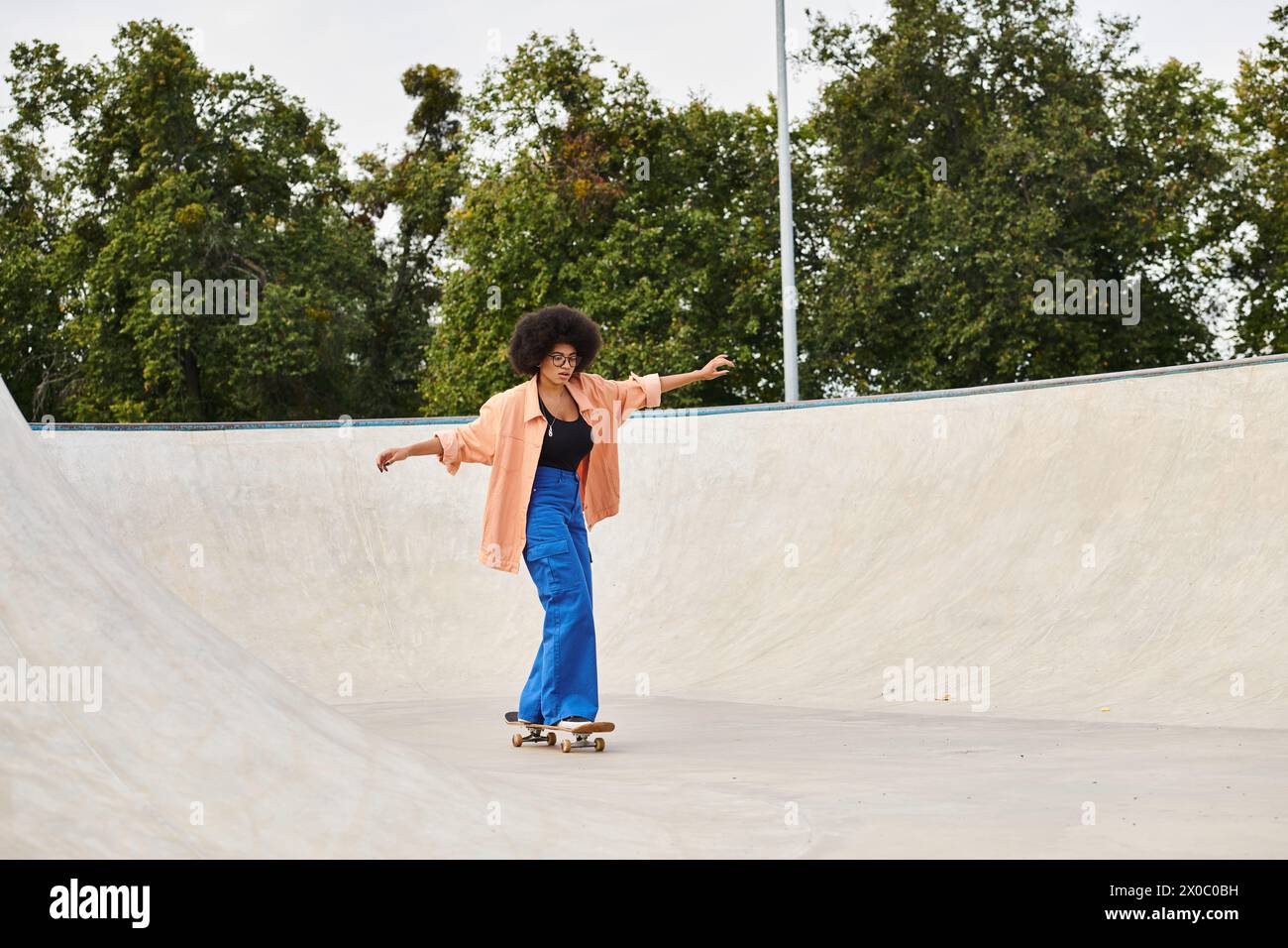 A young African American woman with curly hair confidently rides her skateboard up the side of a ramp in a skate park. Stock Photo