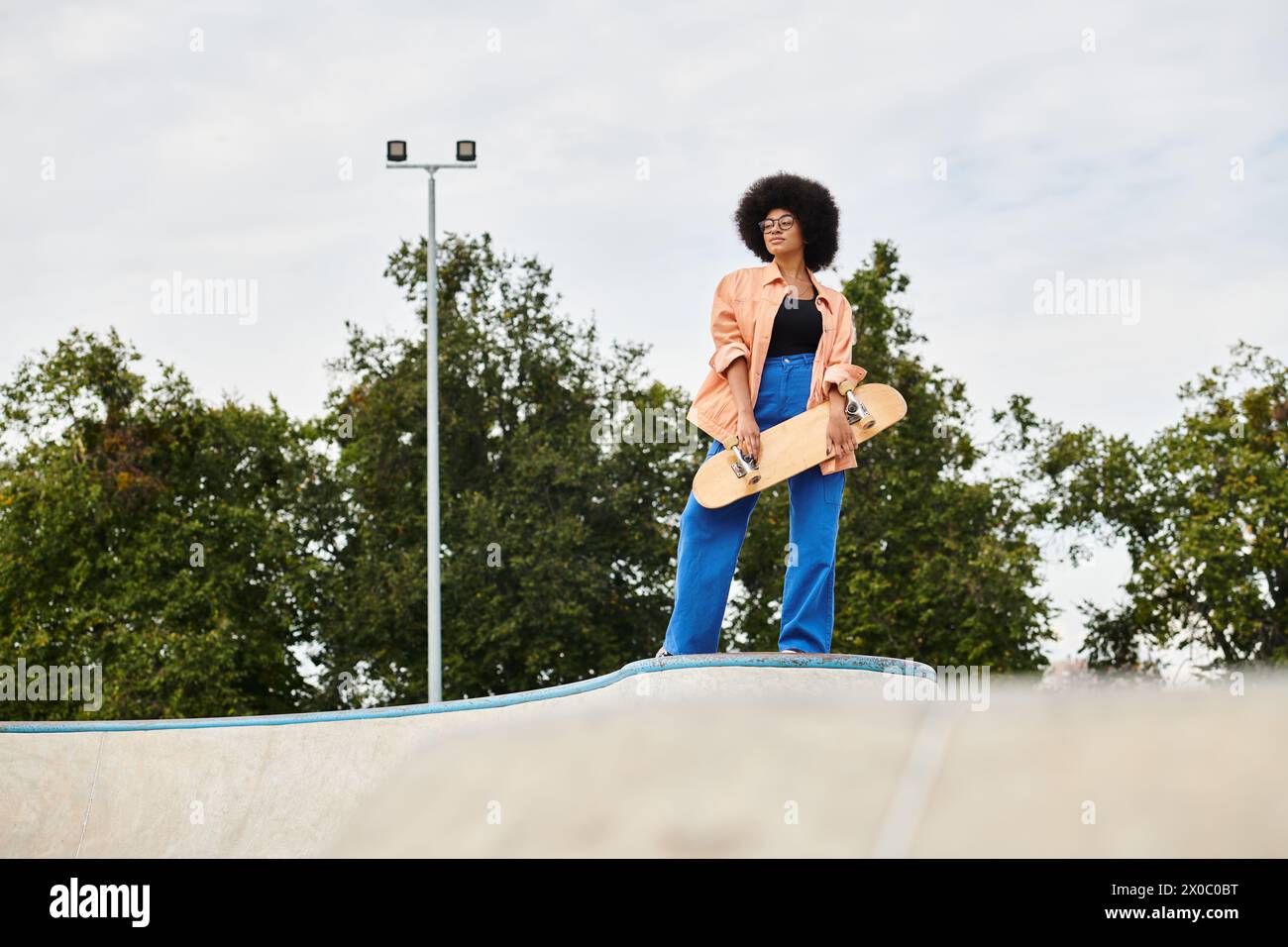 A young African American woman with curly hair confidently stands atop a skateboard ramp at an outdoor skate park. Stock Photo