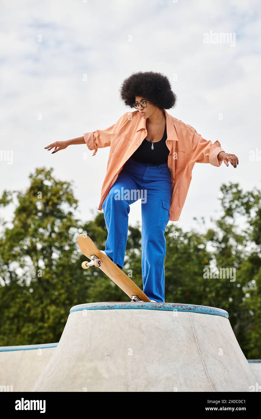 A young African American woman with curly hair rides a skateboard on top of a cement ramp in an outdoor skate park. Stock Photo