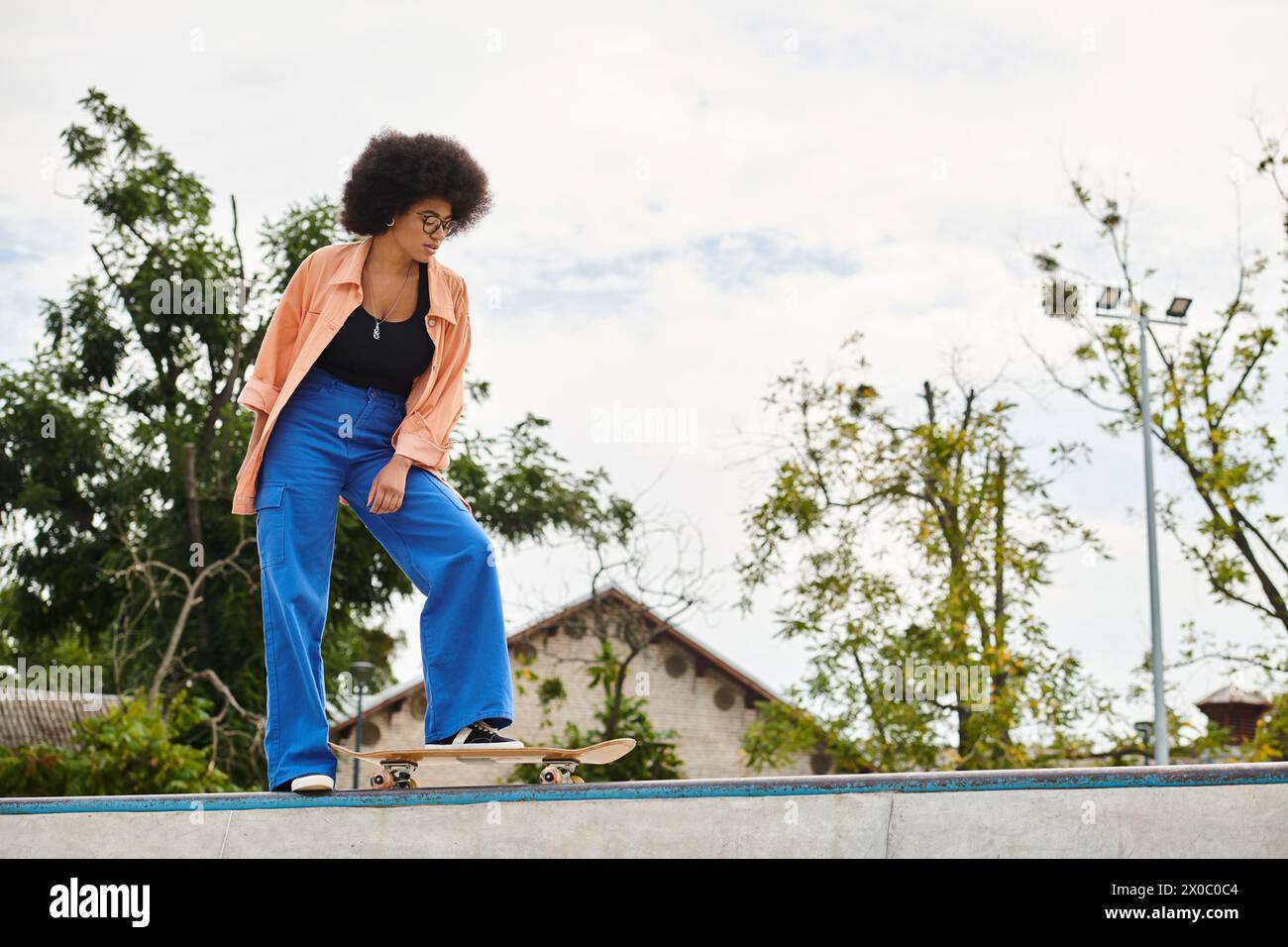 Young African American woman with curly hair riding skateboard on top of cement wall in outdoor skate park. Stock Photo