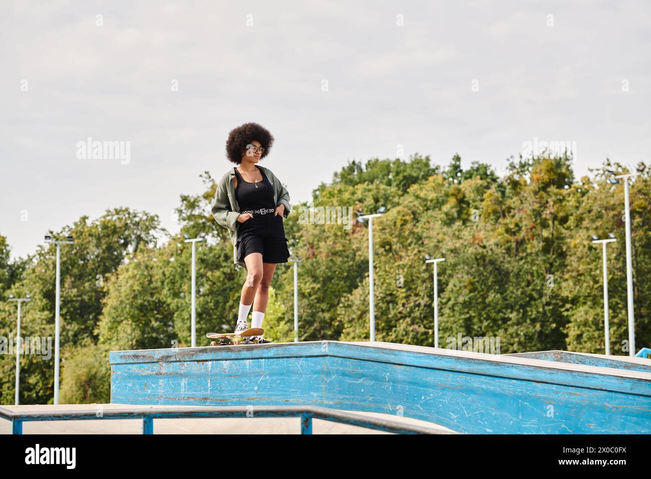 An African American woman with curly hair confidently stands on top of a skateboard ramp in an outdoor skate park. Stock Photo