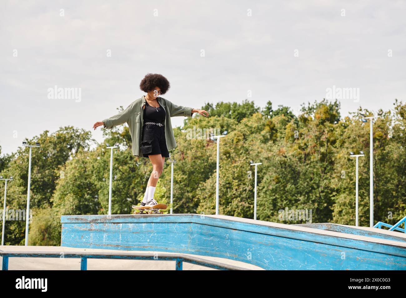 A young African American woman with curly hair skillfully skateboarding on the edge of a pool in an outdoor skate park. Stock Photo