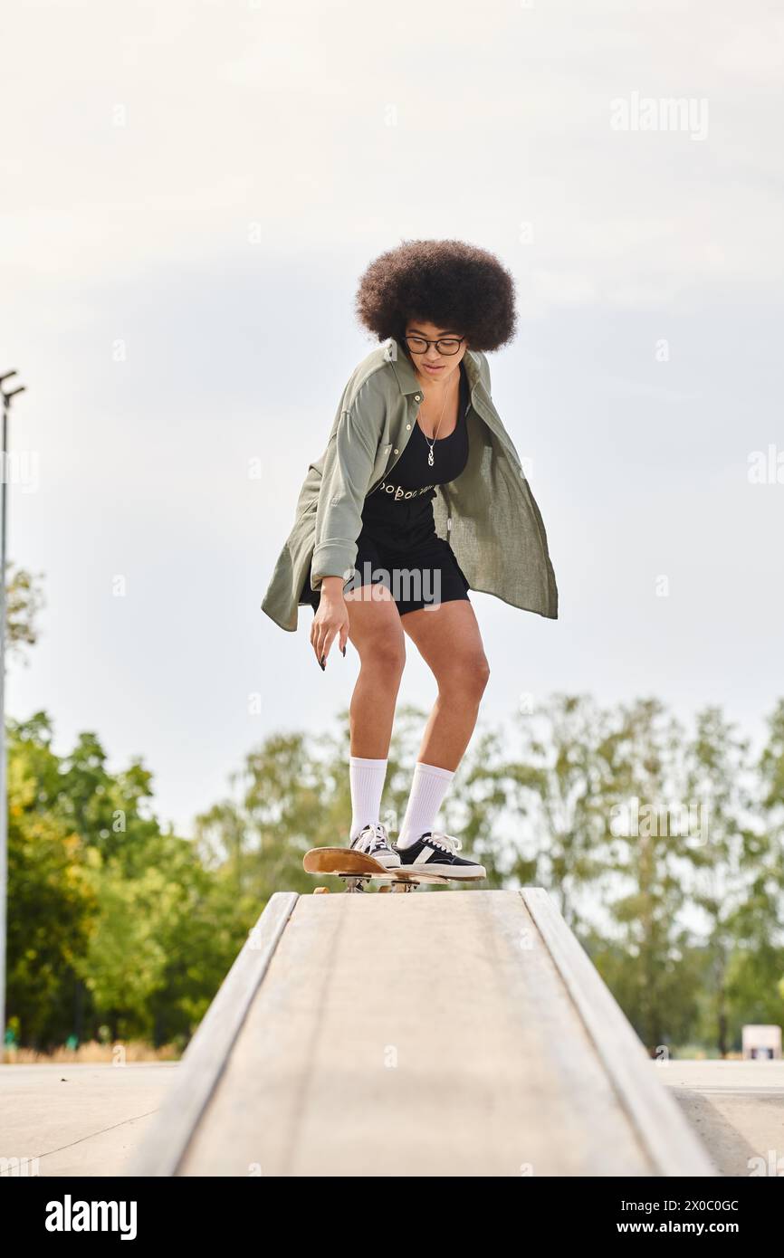 A young African American woman with curly hair skillfully rides a skateboard on a ledge at an urban skate park. Stock Photo