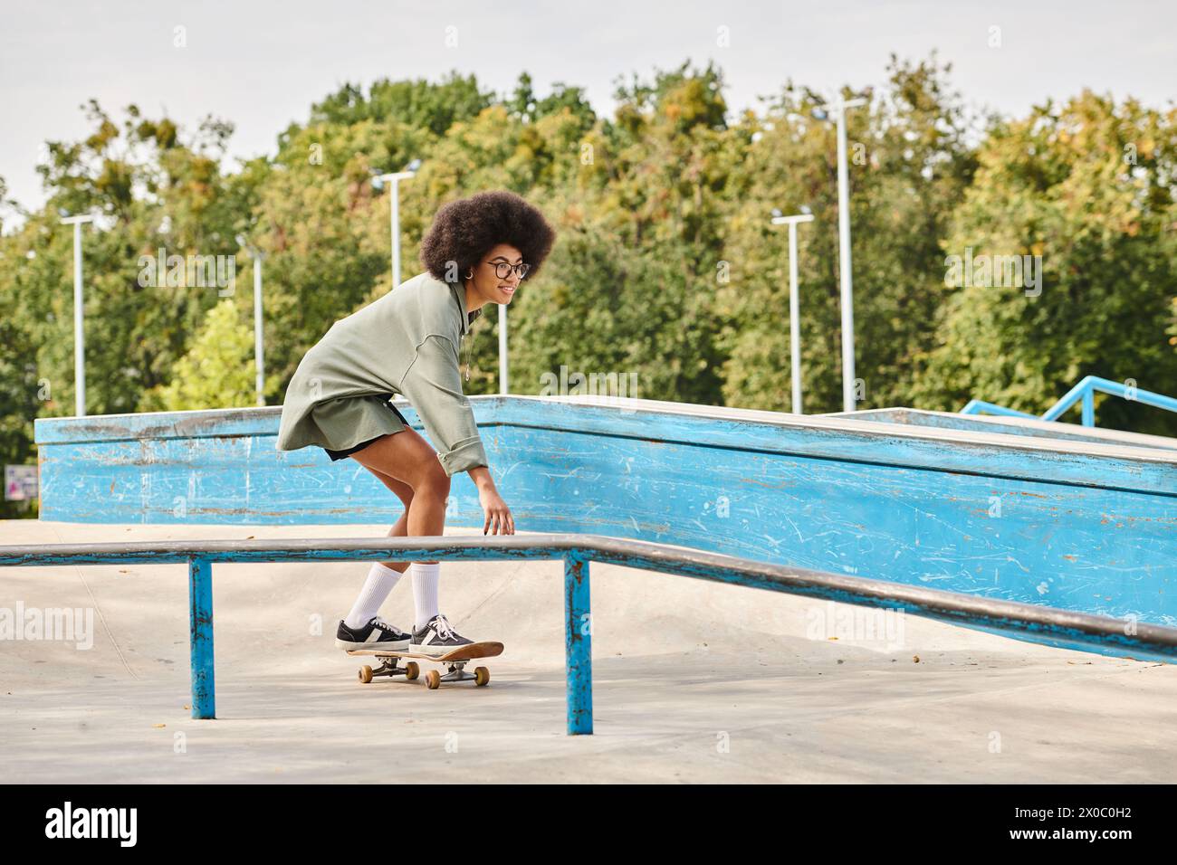 A young African American woman with curly hair skillfully rides a skateboard on a rail in an outdoor skate park. Stock Photo