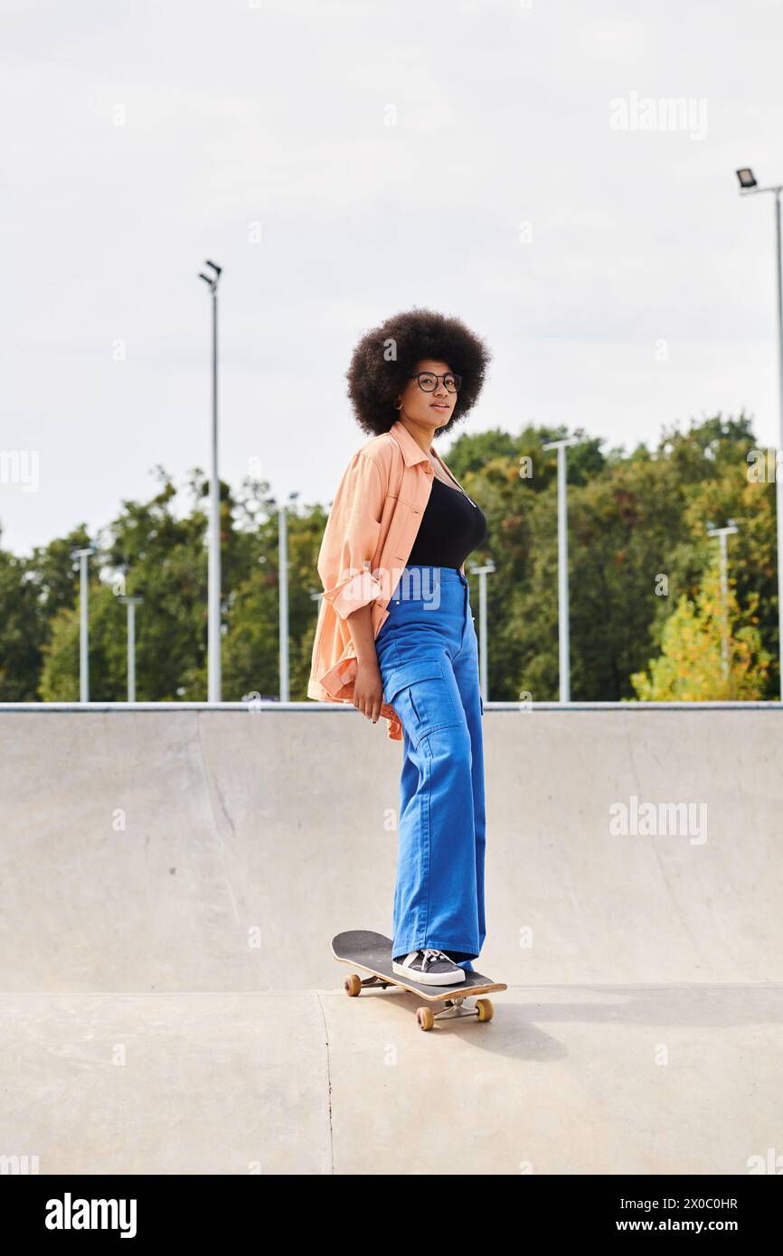 A young African American woman with curly hair confidently stands on a skateboard at a vibrant skate park. Stock Photo