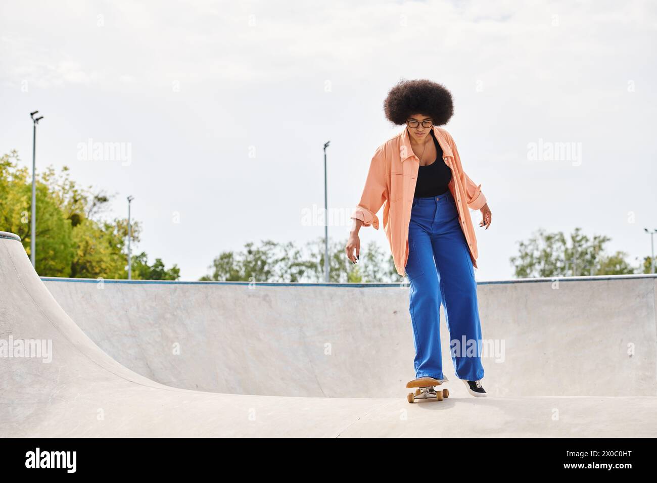 A young African American woman with curly hair skilfully rides a skateboard up the side of a ramp in a vibrant skate park. Stock Photo
