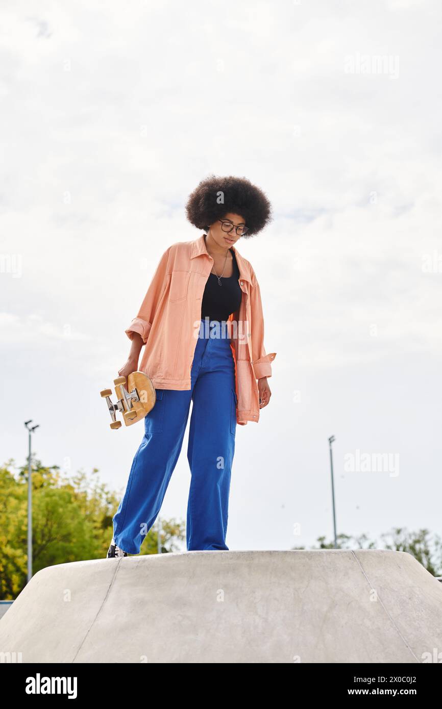 A young African American woman with curly hair confidently skateboards on top of a cement ramp in an outdoor skate park. Stock Photo