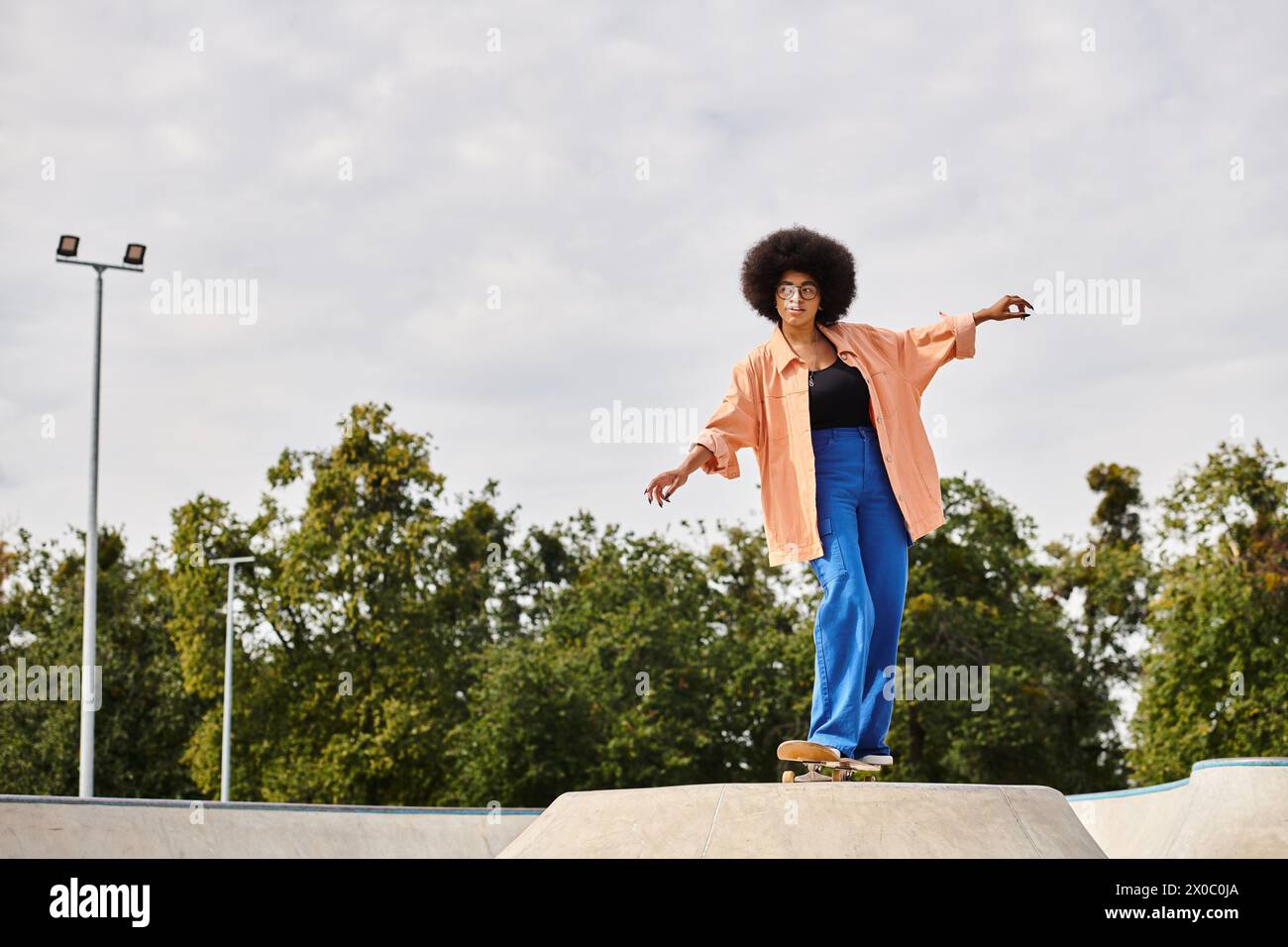 Young African American woman with curly hair skateboarding on top of a cement ramp in a skate park. Stock Photo
