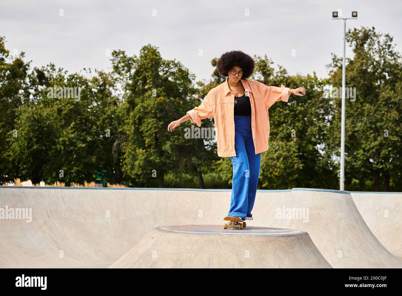 A young African American woman with curly hair confidently rides her skateboard up the side of a ramp at a skate park. Stock Photo