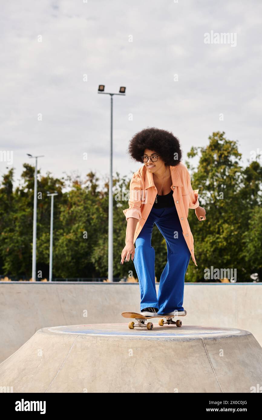 A young African American woman with curly hair rides a skateboard on top of a cement ramp in an urban skate park. Stock Photo