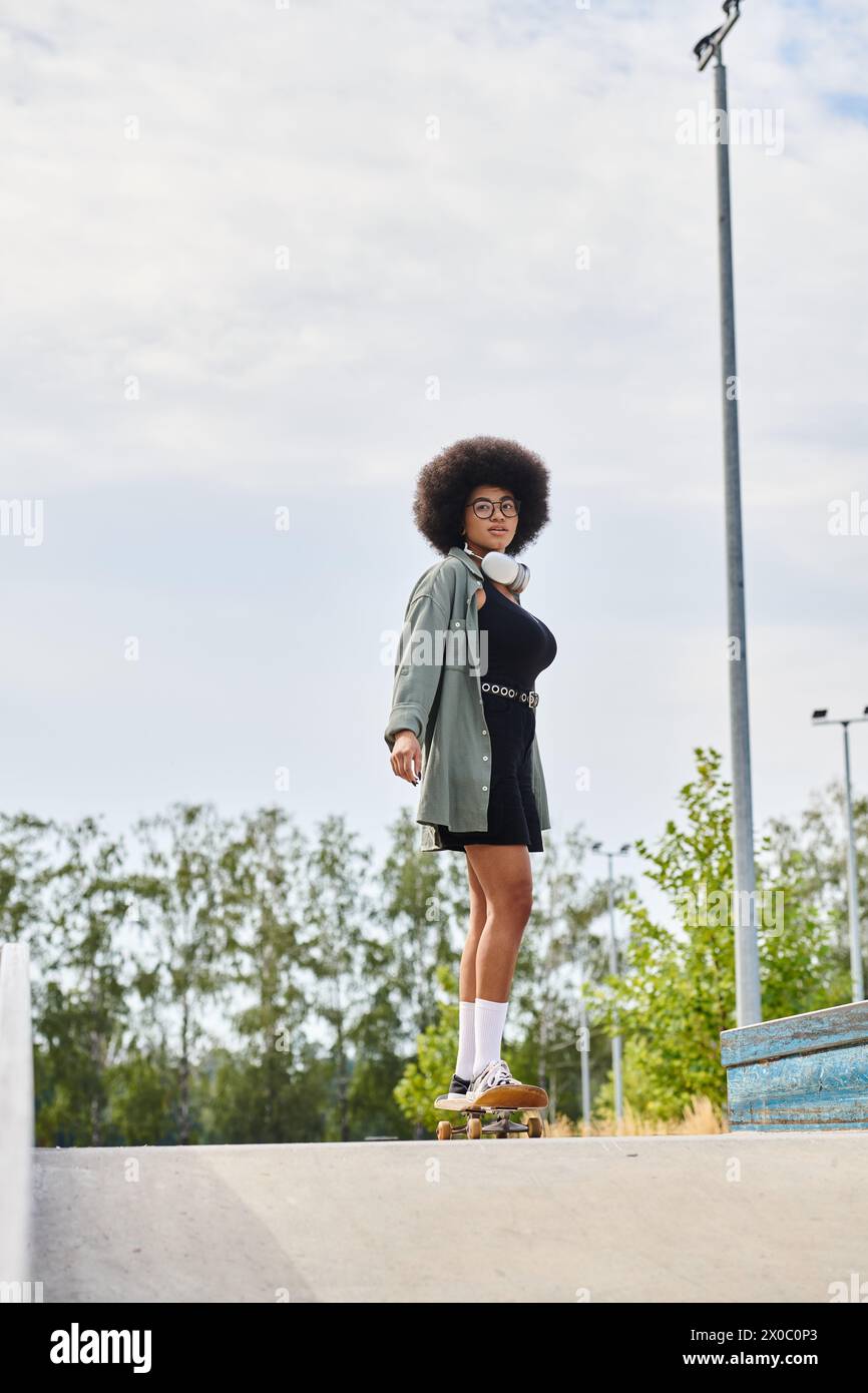 A young African American woman with curly hair skillfully balances on a skateboard at the top of a ramp in a vibrant skate park setting. Stock Photo