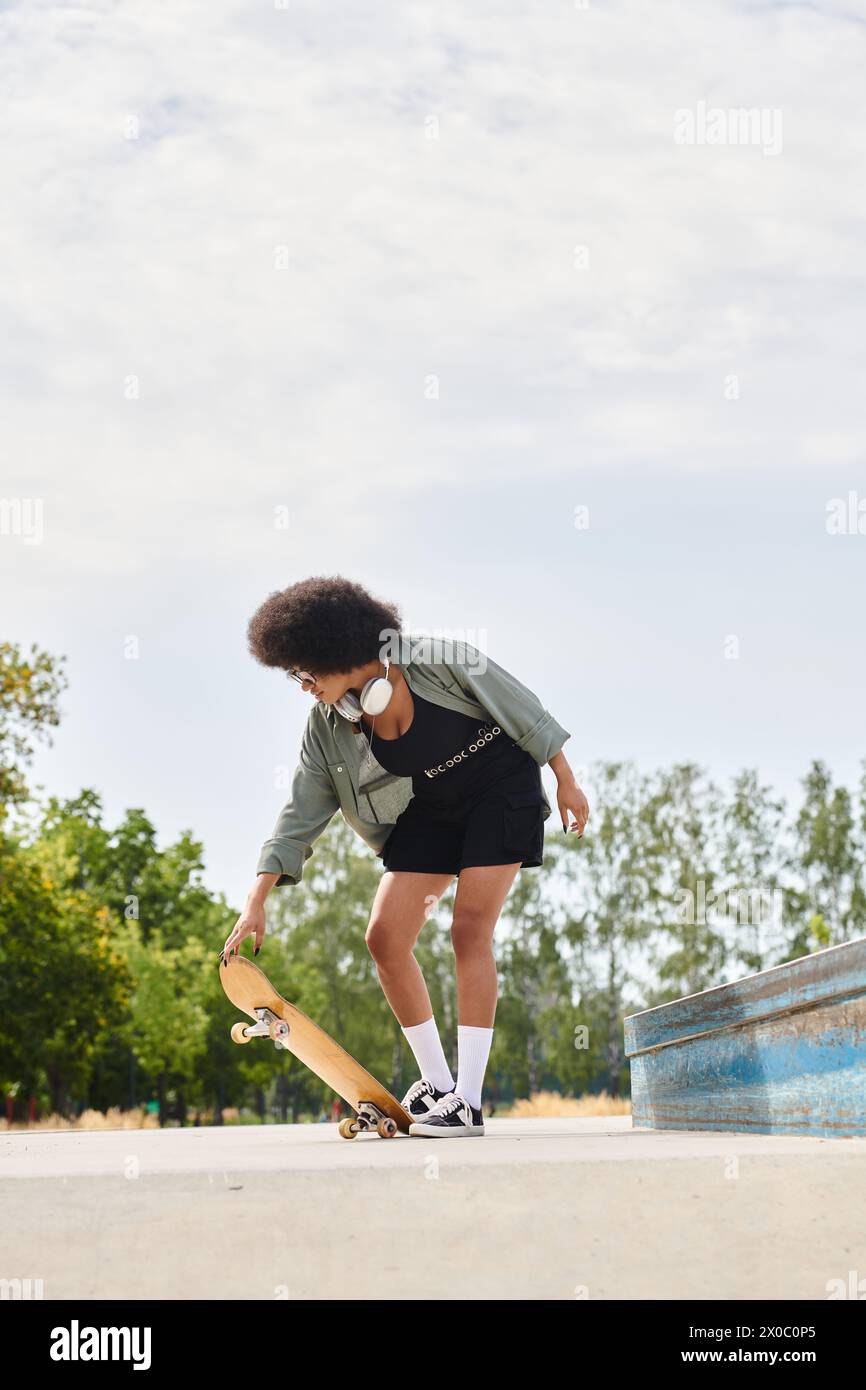 African American woman with curly hair glides on skateboard in a sleek black dress at an outdoor skate park. Stock Photo