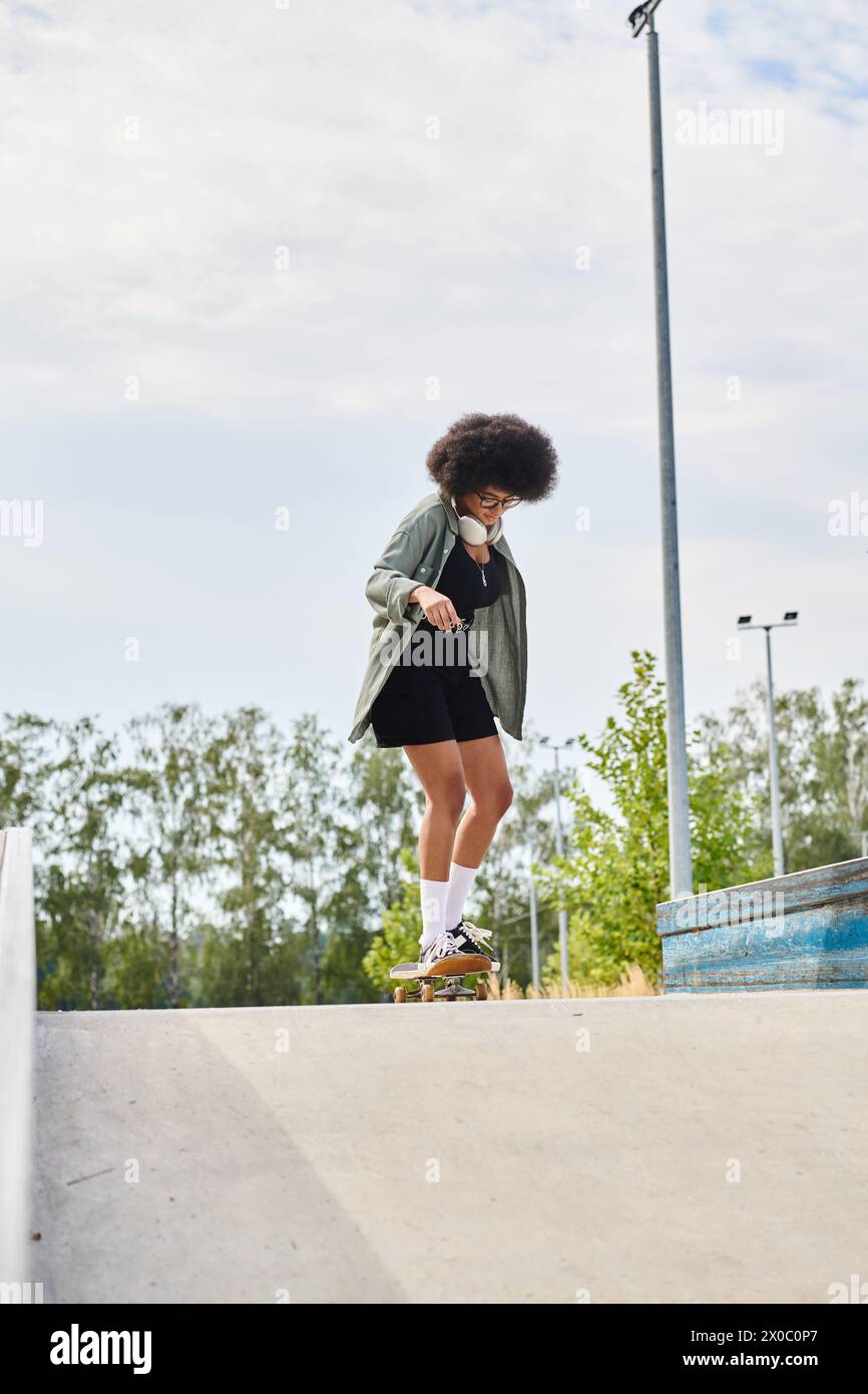 A young African American woman with curly hair confidently rides a skateboard down a cement ramp at an outdoor skate park. Stock Photo