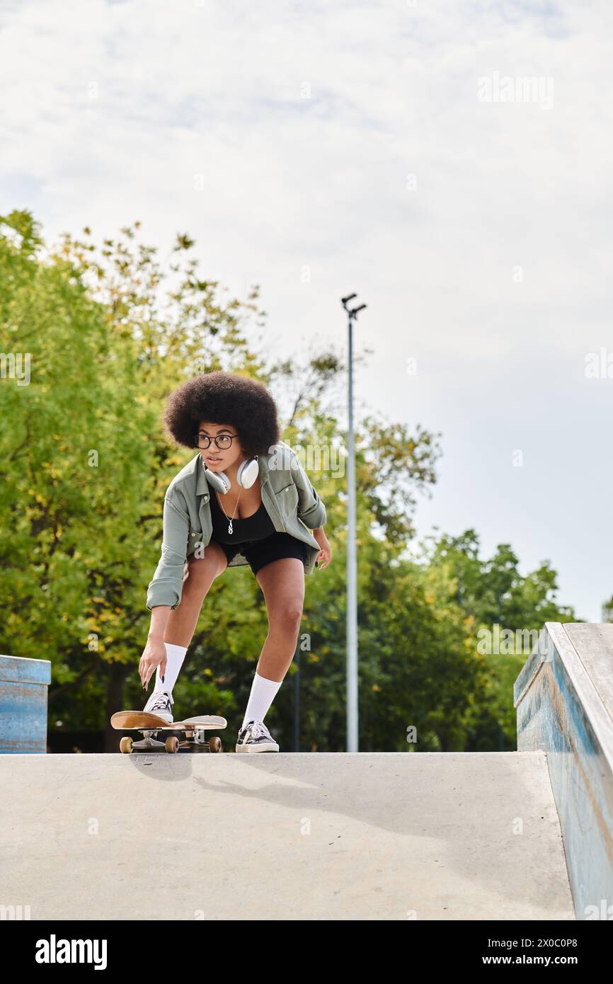 A young African American woman with curly hair skateboard down the side of a ramp at a vibrant outdoor skate park. Stock Photo