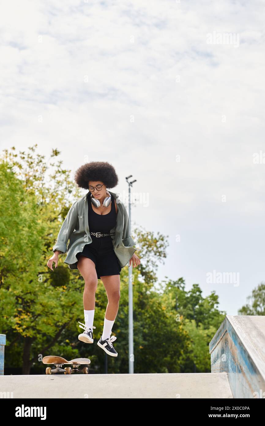 A young African American woman with curly hair jumps her skateboard high in the air at an outdoor skate park. Stock Photo