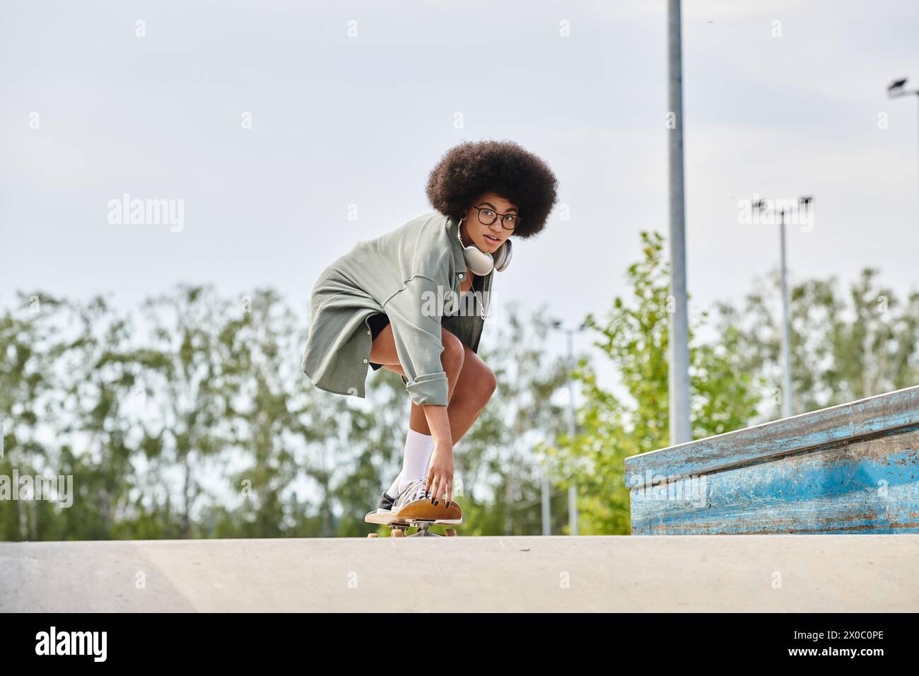 A young African American woman with curly hair confidently rides a skateboard down the ramp in a vibrant skatepark. Stock Photo