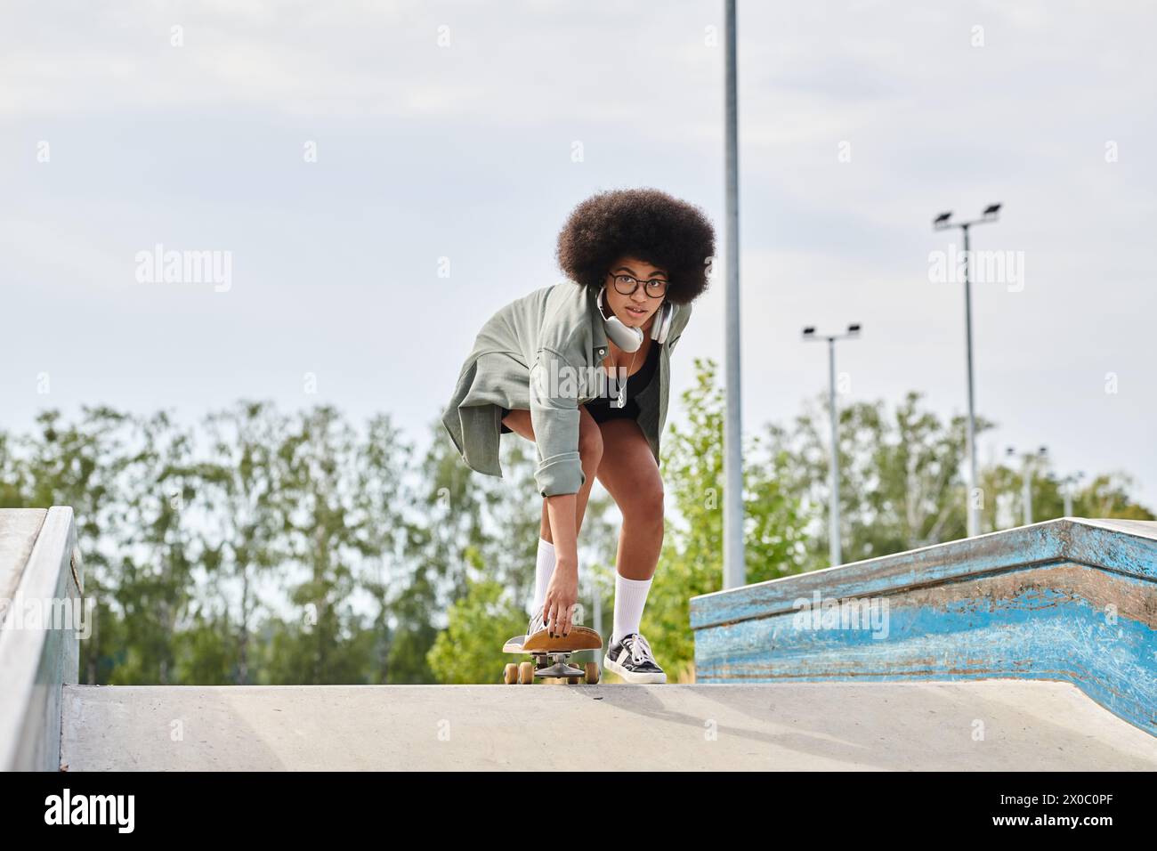 Young African American woman with curly hair skilfully rides a skateboard on a ramp at an outdoor skate park. Stock Photo
