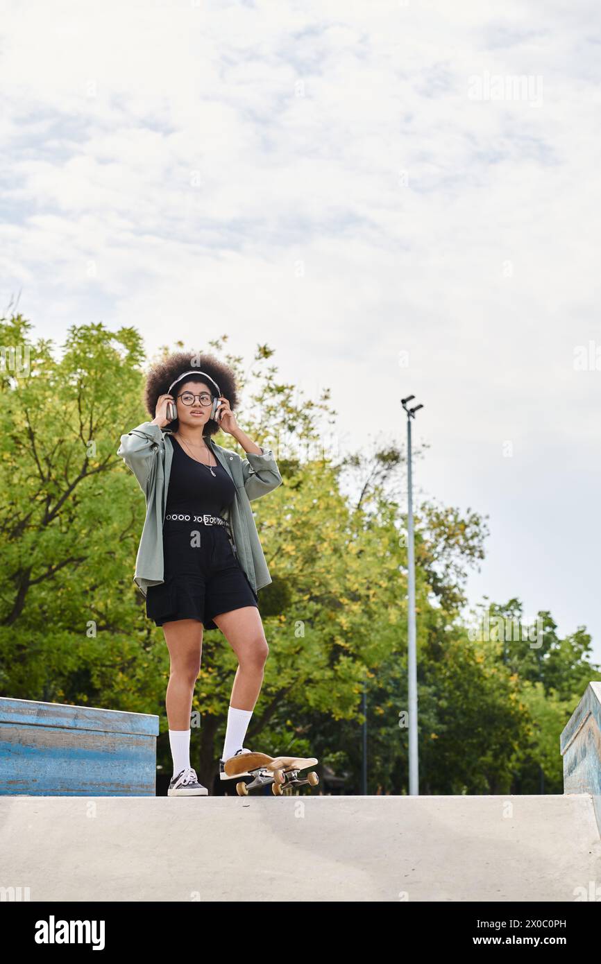 A young African American woman with curly hair confidently stands on top of a skateboard ramp in a skate park. Stock Photo