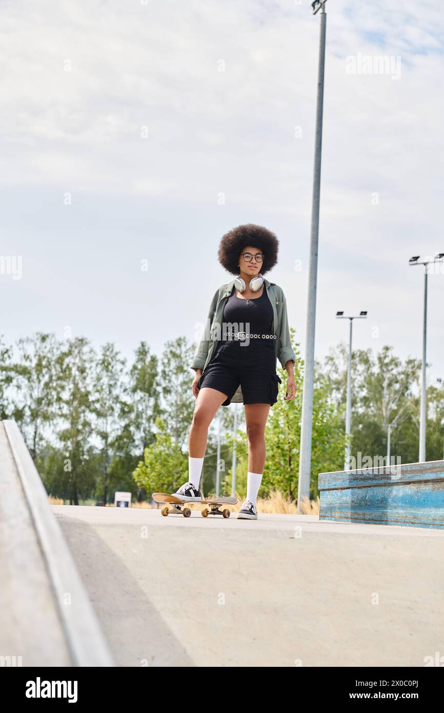 A young African American woman with curly hair confidently skateboards down a city sidewalk on a sunny day. Stock Photo
