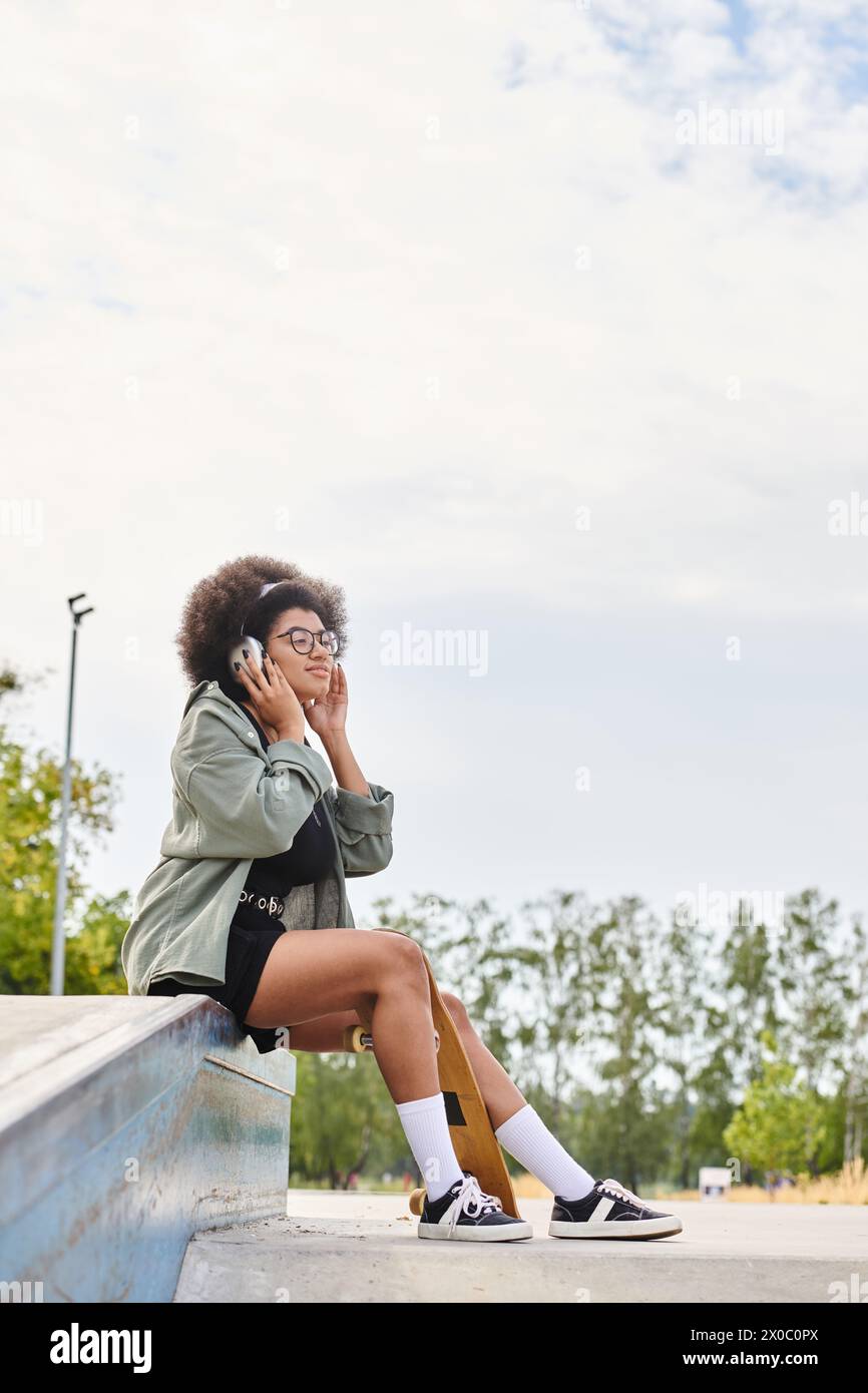 A young African American woman with curly hair, sitting on a ledge, engaged in a phone conversation in an urban setting. Stock Photo