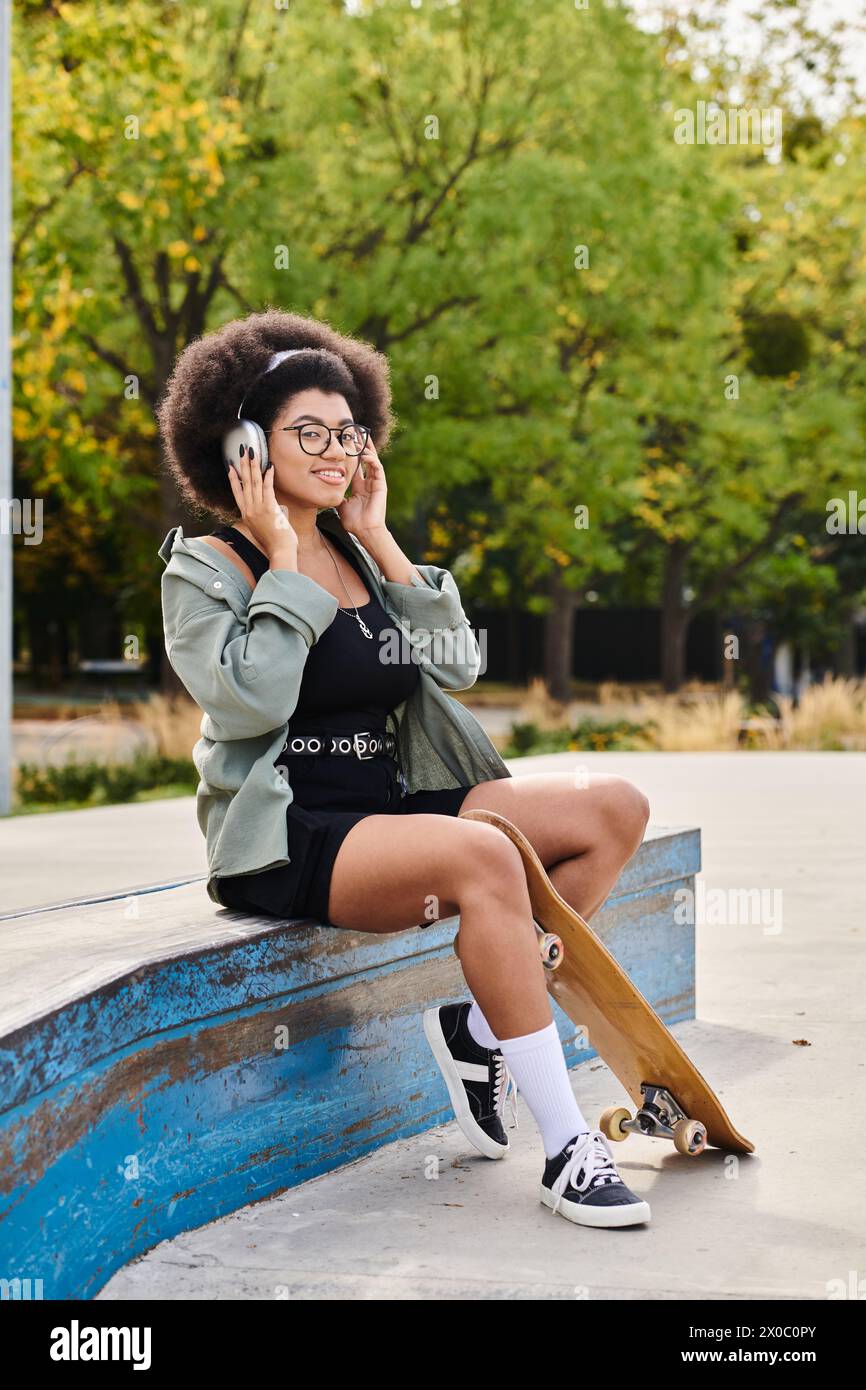 A young African American woman with curly hair sits on a bench, engrossed in a conversation on her cell phone at a skate park. Stock Photo
