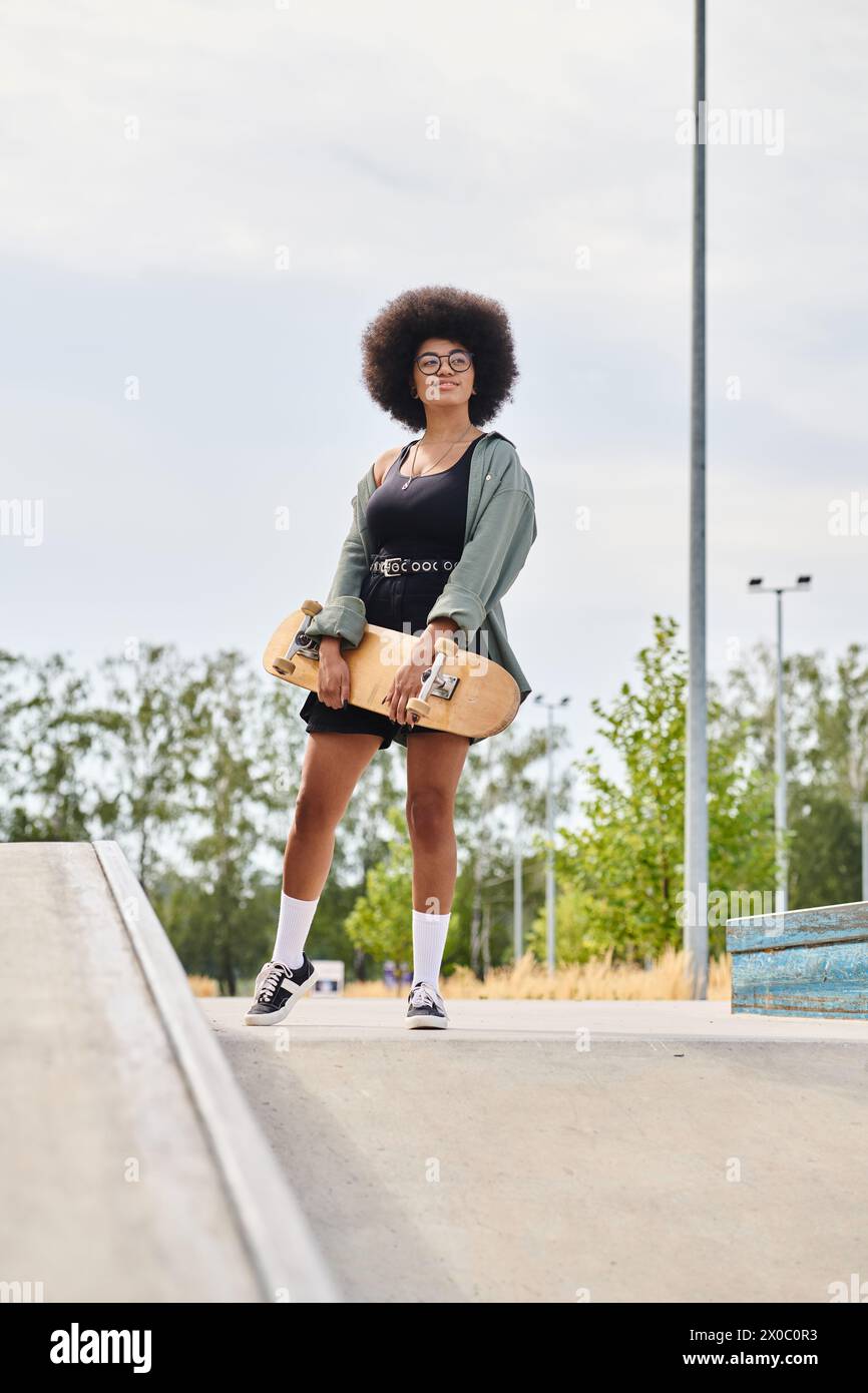 A young African American woman with curly hair confidently stands on a skateboard on a ramp in a skate park. Stock Photo