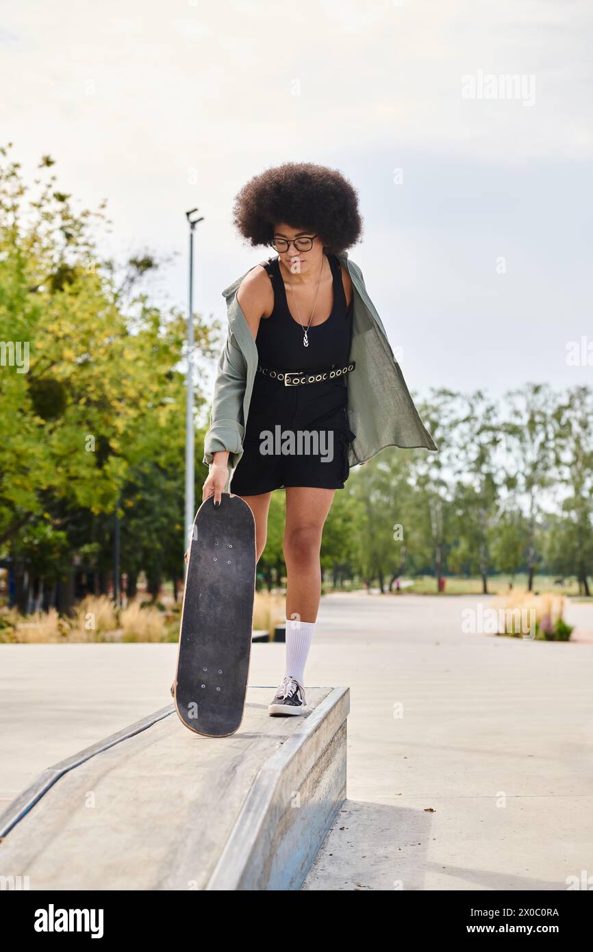 A young African American woman with curly hair exudes style as she gracefully holds a skateboard in a black dress at a skate park. Stock Photo
