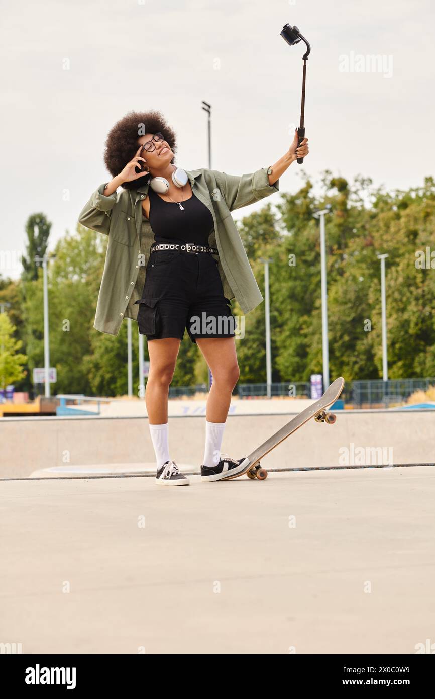 Young African American woman with curly hair skateboarding in a skate park while holding cellphone. Stock Photo
