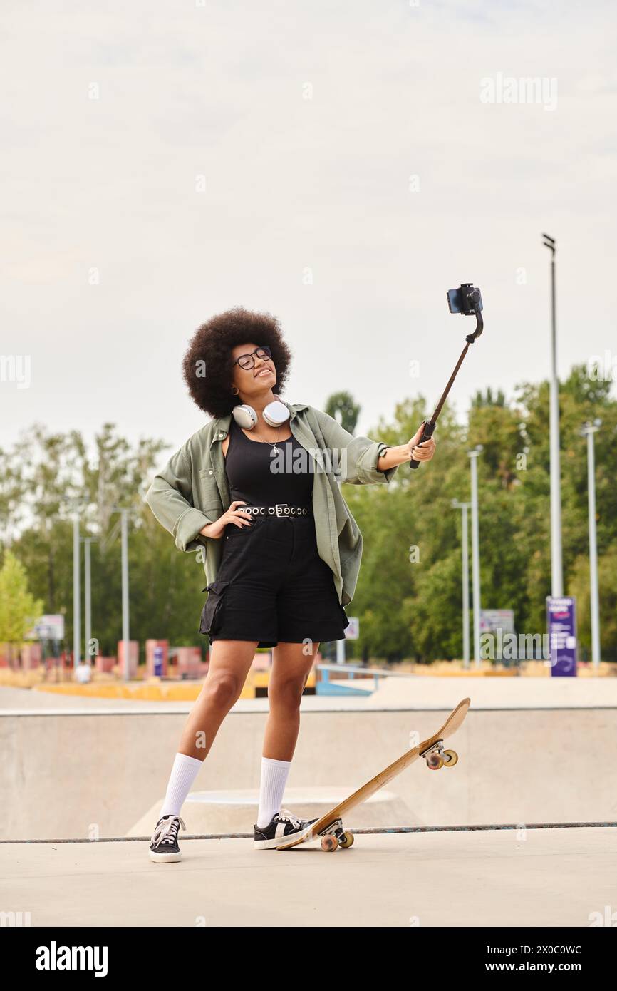 A young African American woman with curly hair stands next to a skateboard, holding a selfie stick in a skate park. Stock Photo