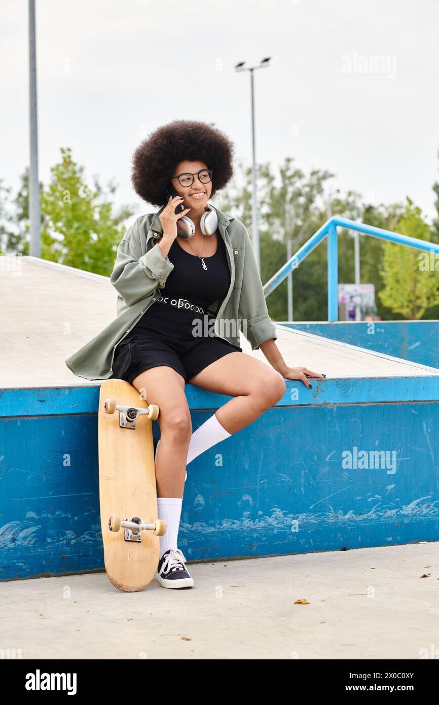 A young African American woman with curly hair sits on a skateboard, talking on a cell phone in a skate park. Stock Photo
