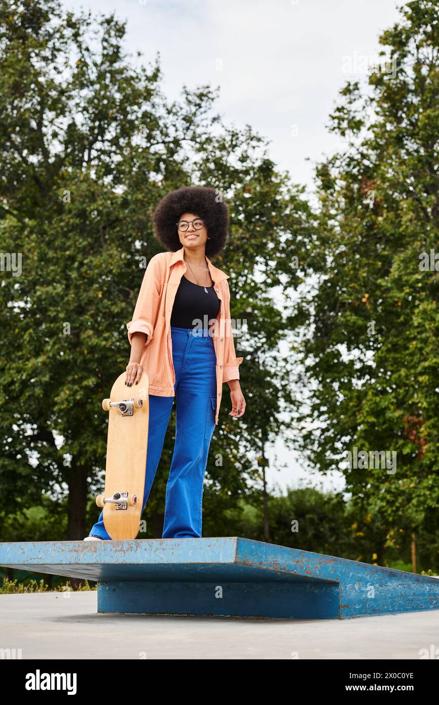 Young African American woman with curly hair holding a skateboard on top of a blue platform at a skate park. Stock Photo