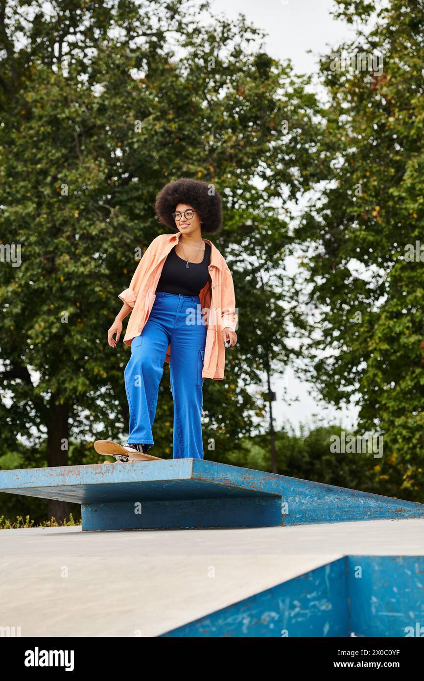 A young African American woman with curly hair gracefully stands on top of a blue object in a skate park. Stock Photo