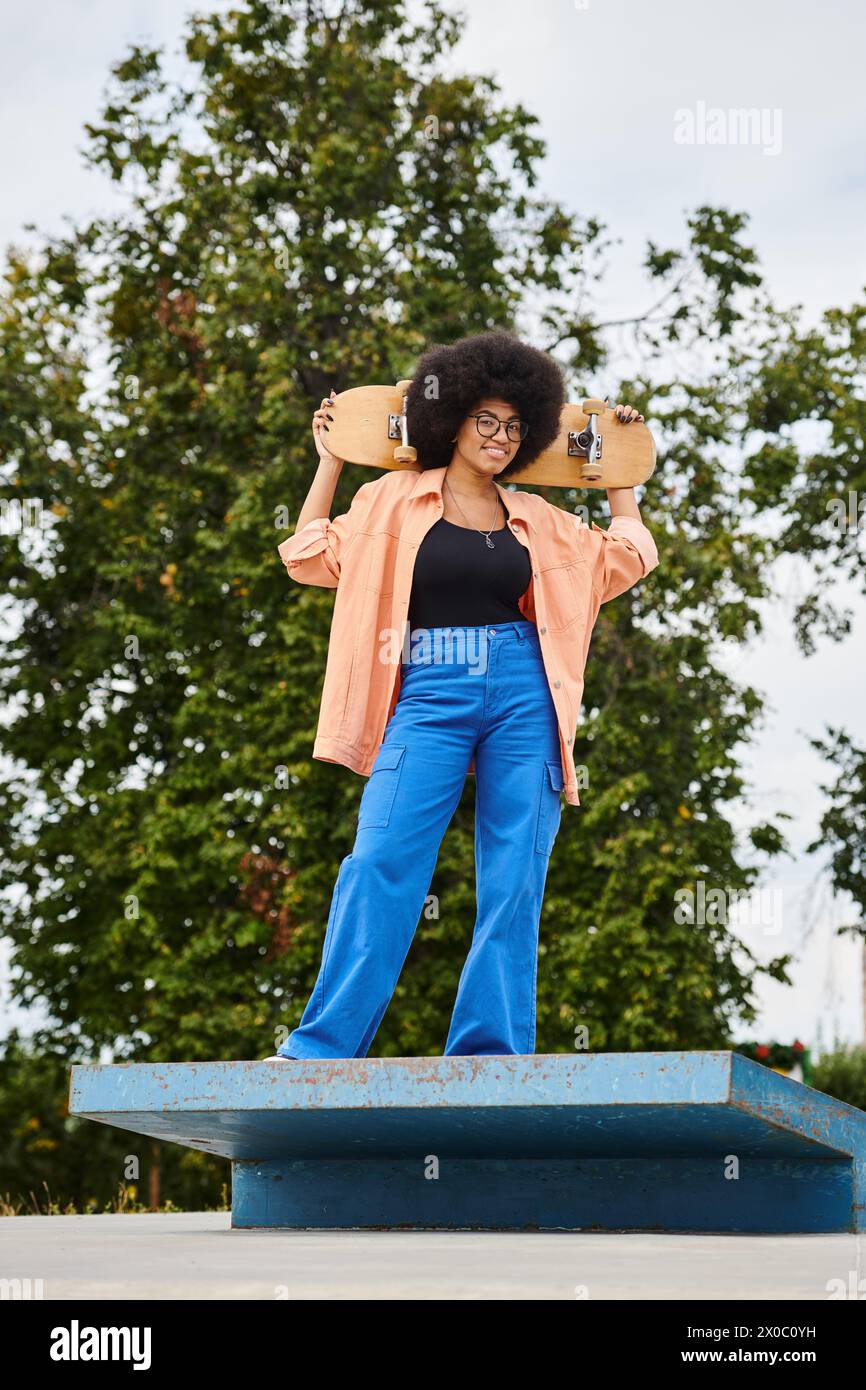 African American woman with curly hair strikes a pose holding a skateboard on a blue platform in a skate park. Stock Photo