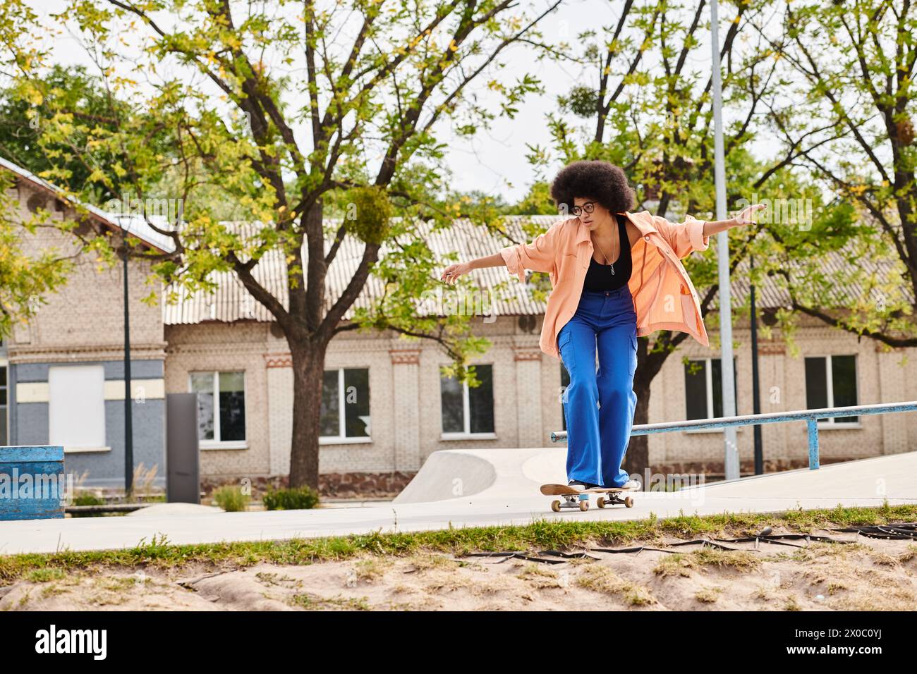 A young African American woman with curly hair gracefully rides a skateboard down a sidewalk in a skate park. Stock Photo